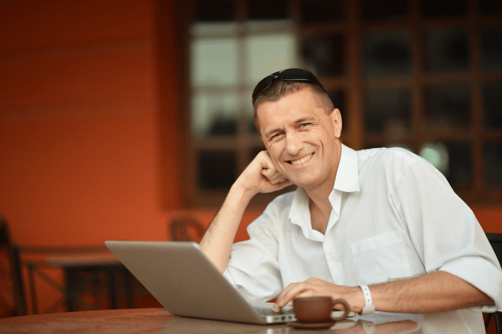 smiling man with eyes that aren't dry, sitting in front of a laptop with a cup of coffee - there's a rust-colored wall behind him